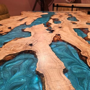 May include: A close-up of a wooden table with a unique design. The table features a light brown wood grain with sections filled with a vibrant, swirling teal-colored resin. The contrast between the wood and resin creates a striking visual effect.