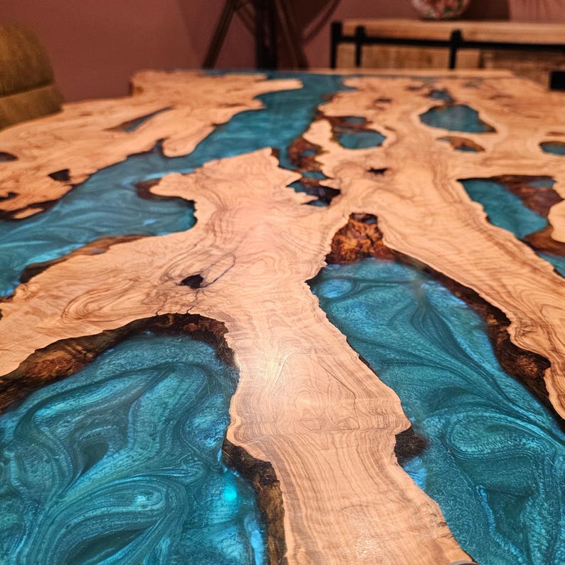 May include: A close-up of a wooden table with a unique design. The table features a light brown wood grain with sections filled with a vibrant, swirling teal-colored resin. The contrast between the wood and resin creates a striking visual effect.