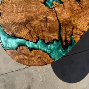 May include: Close-up of a wooden table top with a river of teal-colored resin. The wood grain is a rich brown with intricate patterns. The resin flows through the wood, creating a unique and artistic design.