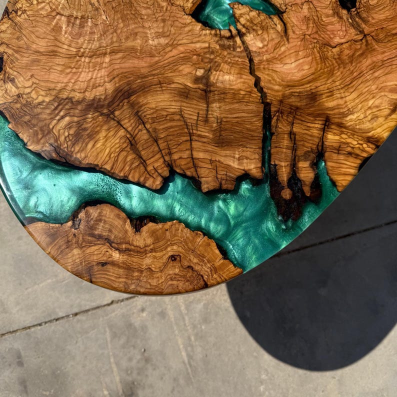 May include: Close-up of a wooden table top with a river of teal-colored resin. The wood grain is a rich brown with intricate patterns. The resin flows through the wood, creating a unique and artistic design.