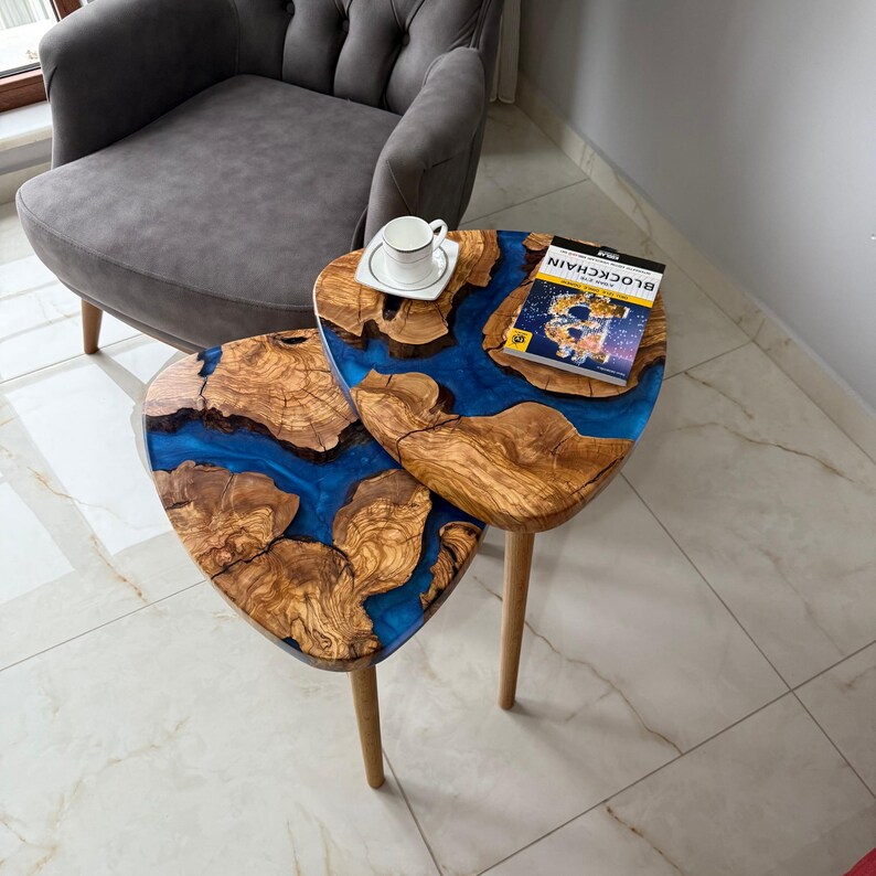 May include: Two wooden side tables with a unique design. The tables feature a blue resin river flowing through the natural wood grain. One table has a white teacup and saucer, the other has a book.
