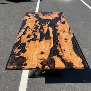 May include: A rectangular wooden table with a unique design. The tabletop features a light brown wood grain pattern with dark brown epoxy resin filling the spaces. The table is supported by a black metal base. The table measures approximately 72 inches long.