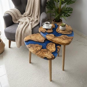 Op de afbeelding: Twee houten bijzettafels met een blauw hars rivierontwerp. De tafels hebben een natuurlijk houtnerfpatroon en worden ondersteund door drie lichtgekleurde houten poten. Een kopje koffie en een kleine plant staan erop.