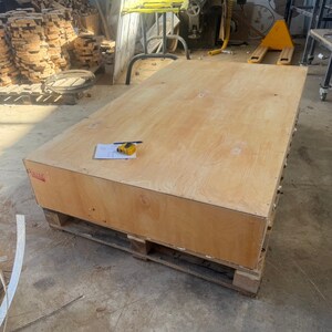 May include: A large, rectangular wooden crate made of light-colored plywood sits on a wooden pallet. A sheet of paper, a pen, and a yellow measuring tape are on top of the crate. The crate is in a workshop setting.