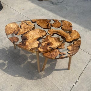 Può includere: Un set di due tavolini da caffè con piano in resina trasparente e intarsi in legno naturale. I tavoli hanno gambe in legno di colore chiaro. Il tavolo più grande è lungo circa 100 cm, e il più piccolo circa 50 cm.