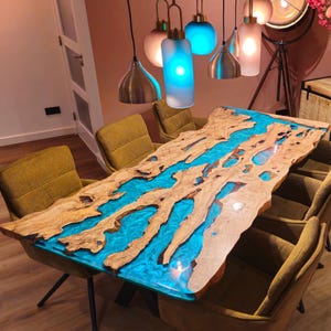 May include: A handcrafted wooden dining table with a river of blue resin. The table has a natural edge and is surrounded by yellow-green upholstered chairs. Decorative pendant lights hang above the table, illuminating the room.