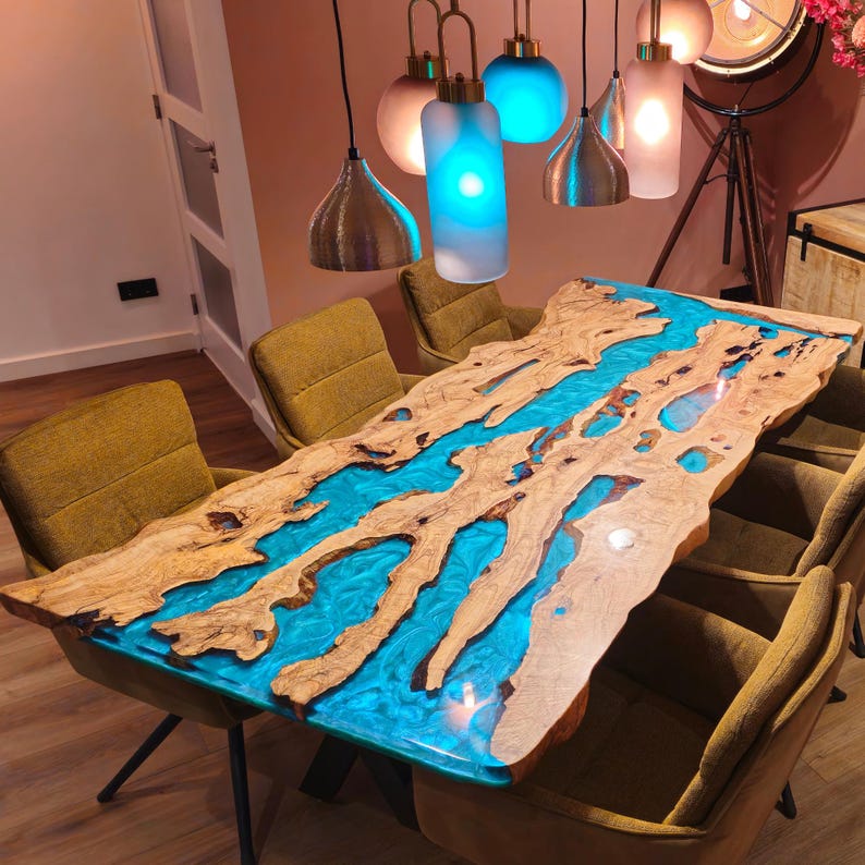 May include: A handcrafted wooden dining table with a river of blue resin. The table has a natural edge and is surrounded by yellow-green upholstered chairs. Decorative pendant lights hang above the table, illuminating the room.