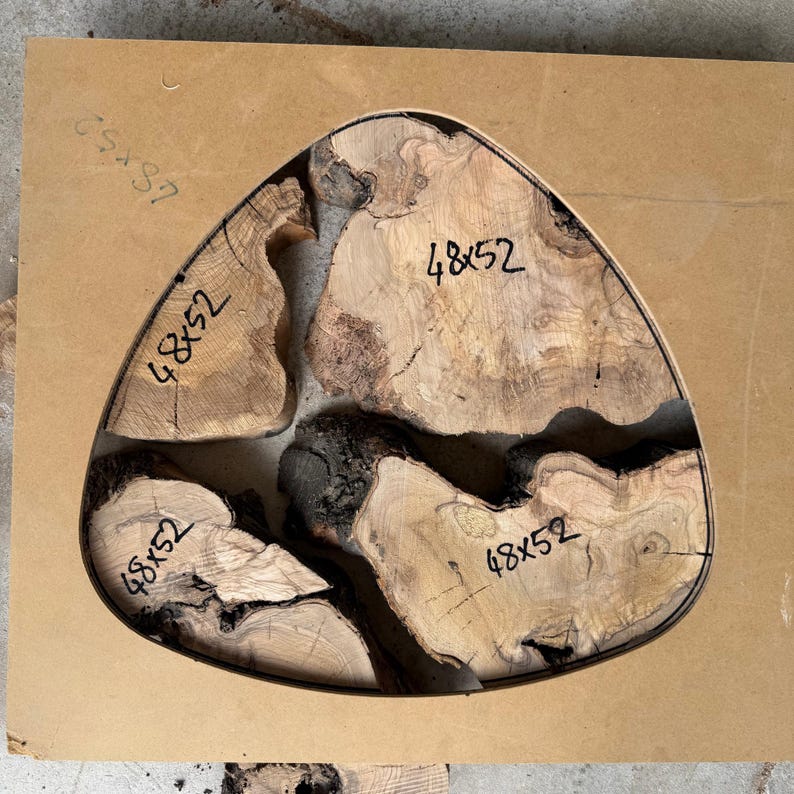 May include: A wooden, triangular table top with a live edge design. The wood is light brown with dark brown and black accents. The dimensions are 48 x 52 inches. The table top is set on a light brown surface.