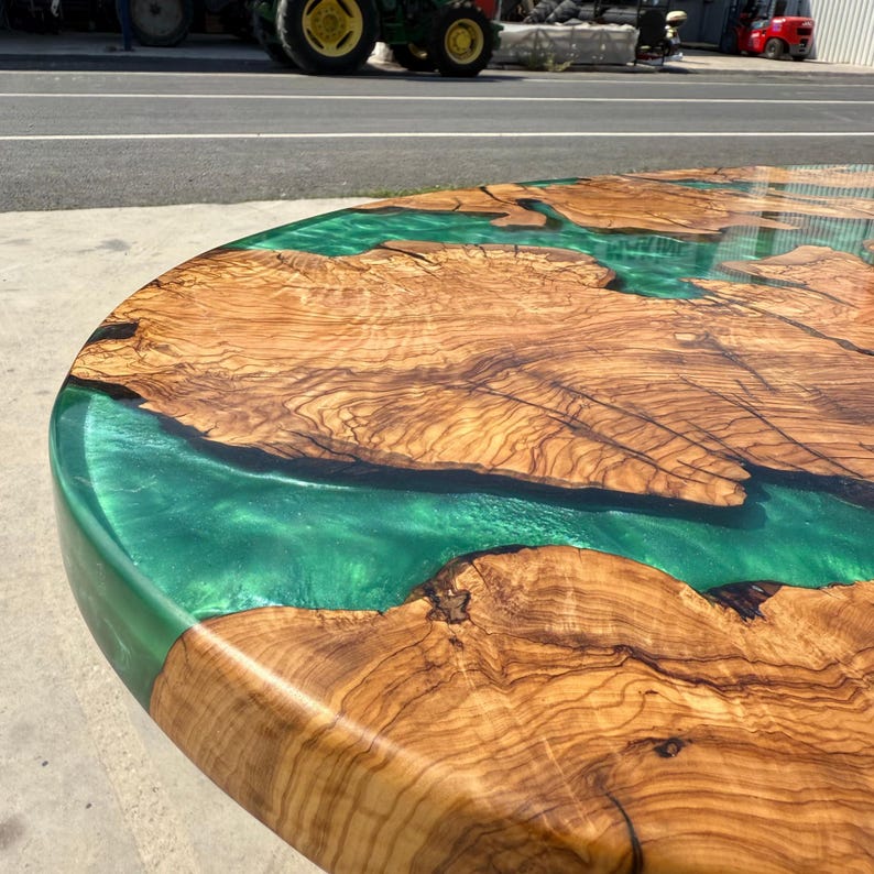 May include: A round wooden table with a river of green epoxy resin running through the center. The wood has a natural grain pattern with shades of brown and tan. The table's edge is also green, creating a cohesive design.