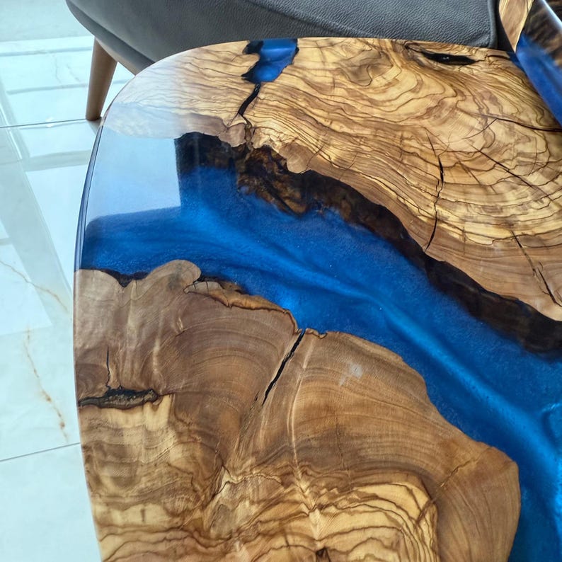 May include: Close-up of a wooden table with a river of blue resin. The table's surface is made of light brown wood with visible grain and cracks. The blue resin flows through the wood, creating a unique design. The table has a glossy finish.