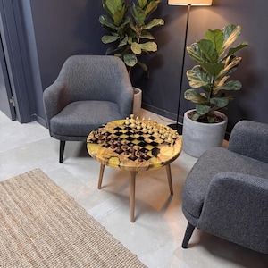 May include: A round wooden chess table with a black and yellow resin top, featuring a chess set in play. The table has three wooden legs and is surrounded by gray armchairs and potted plants.