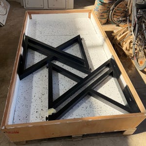 May include: Black metal table legs in a wooden crate. The legs are in a geometric design, with a triangular shape. The crate has a white speckled interior. The legs are likely for a table or desk.