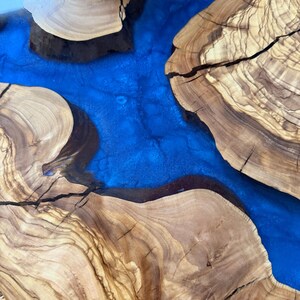 May include: A close-up view of a wooden table with a river of blue resin. The wood has a natural grain pattern with shades of brown and tan. The blue resin flows between the wood sections, creating a striking contrast.