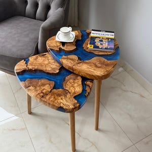 May include: Two-tiered side table with a unique design. The tabletop features a combination of natural wood and blue resin, creating a striking contrast. The table has three wooden legs. A cup and saucer and a book are on the table.