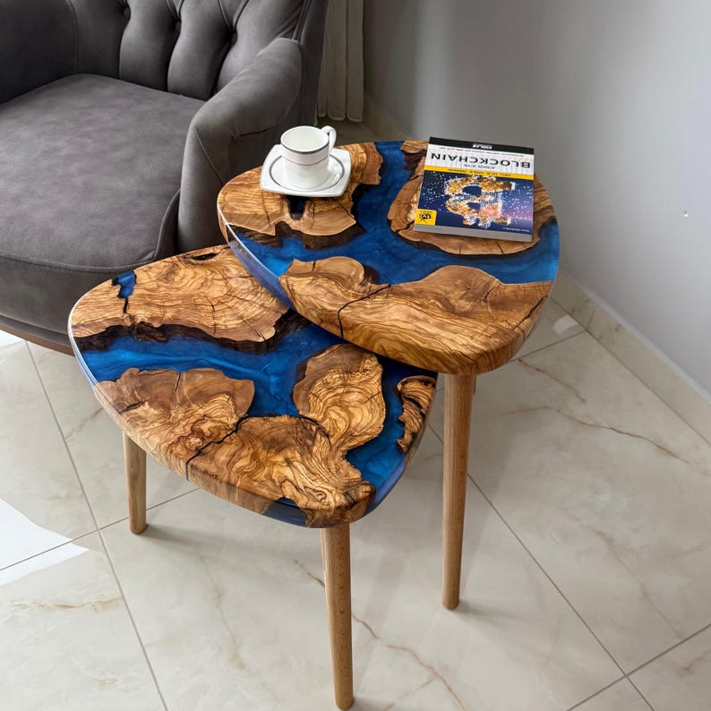 May include: Two-tiered side table with a unique design. The tabletop features a combination of natural wood and blue resin, creating a striking contrast. The table has three wooden legs. A cup and saucer and a book are on the table.
