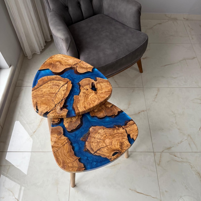 May include: Two wooden side tables with a unique design. The tables feature a natural wood grain pattern with a vibrant blue resin filling the gaps. The tables have three wooden legs and are placed near a gray armchair.