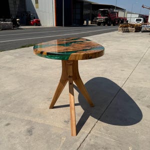 May include: A small, wooden side table with a unique design. The tabletop is a mix of natural wood and green resin, supported by three angled wooden legs. The table is approximately 24 inches tall.