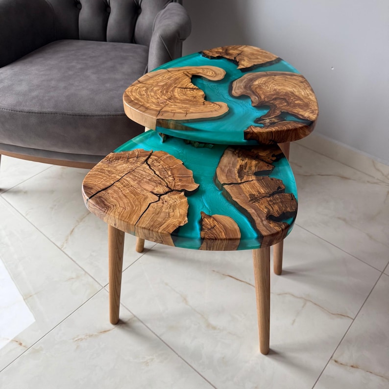 May include: Two wooden side tables with a unique design. The tabletops feature a combination of natural wood and turquoise resin, creating a striking contrast. Each table has three wooden legs. The tables are approximately 20 inches in height.