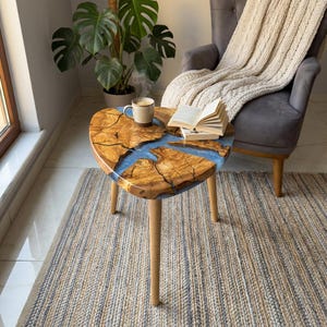May include: A heart-shaped wooden side table with a blue resin river design. The table has three wooden legs and is topped with a cup of coffee and an open book. The table is approximately 24 inches wide.
