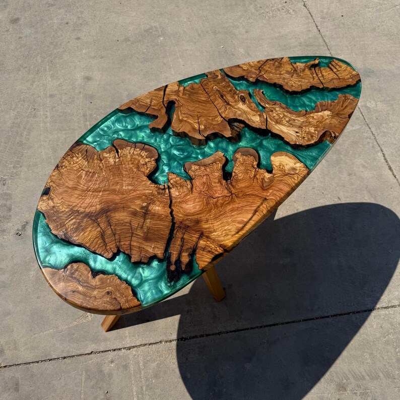 May include: Oval-shaped wooden coffee table with a teal epoxy resin river design. The table features natural wood grain patterns and a glossy finish. The legs are made of light-colored wood. The table is approximately 48 inches long.