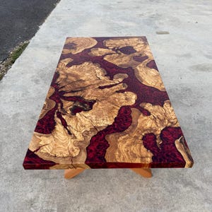 May include: A rectangular wooden coffee table with a unique design. The table features a light brown wood grain pattern with a deep red resin river flowing through the center. The table is supported by light brown wooden legs.