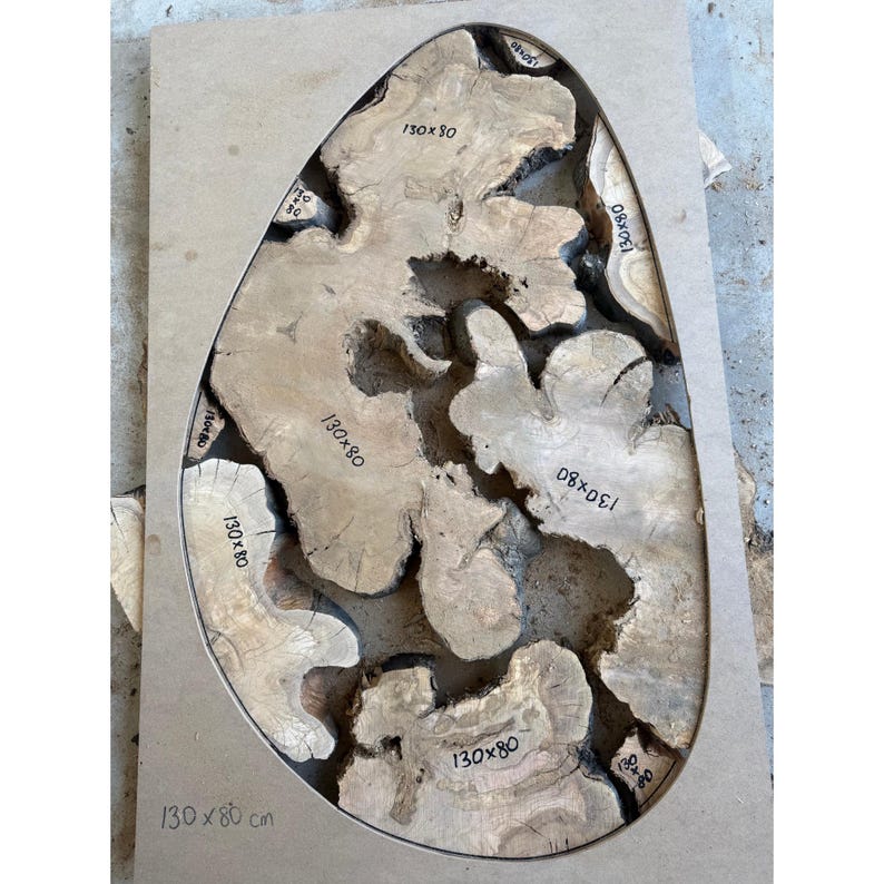 May include: A wooden table top with a unique, organic shape. The surface is made of light brown wood with a dark, resin-filled center. The dimensions are 130 x 80 cm, as indicated on the surface. The design features irregular, flowing shapes.