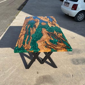 May include: A rectangular wooden table with a unique design. The tabletop features a combination of natural wood grain and a vibrant green epoxy resin. The table is supported by black metal legs, creating a modern aesthetic.