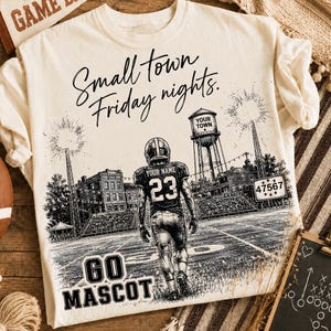 May include: A cream-colored t-shirt featuring a black graphic of a football player on a field with the text Small town Friday nights, YOUR NAME, 23, YOUR TOWN, 47567, and GO MASCOT. The design includes a water tower and stadium lights.