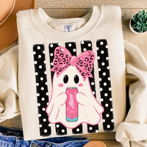 May include: A cream-colored crewneck sweatshirt featuring a graphic of a white ghost wearing a pink leopard print bow. The ghost holds a pink Alani energy drink can. The design is set against a background of vertical black stripes with white polka dots.