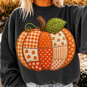 Puede incluir: Una sudadera de cuello redondo gris carbón que presenta un gran gráfico central de una calabaza. La calabaza está diseñada con un patrón de retazos de telas naranjas y blancas, incluyendo cuadros vichy, lunares y estampados florales, con una hoja verde y un tallo marrón.
