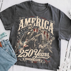 May include: Dark gray t-shirt featuring a graphic of Abraham Lincoln and George Washington playing guitars with the American flag design. The text reads "AMERICA 250 Years 1776-2026".