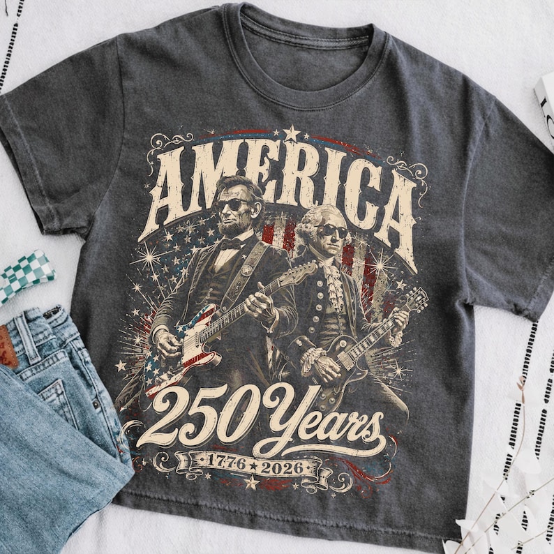 May include: Dark gray t-shirt featuring a graphic of Abraham Lincoln and George Washington playing guitars with the American flag design. The text reads "AMERICA 250 Years 1776-2026".