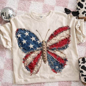 Op de afbeelding: Crèmekleurig T-shirt met een groot vlinderontwerp. De vlindervleugels zijn gemaakt van denim en rood-witte stof, die op de Amerikaanse vlag lijken. Het shirt ligt op een roze en wit geruit oppervlak.