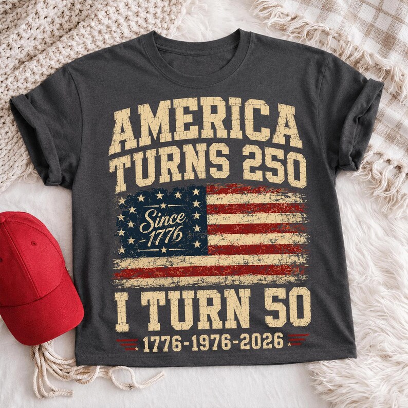 May include: Dark gray t-shirt with the text "AMERICA TURNS 250" above a distressed American flag graphic. Below the flag, it reads "I TURN 50" with the dates 1776-1976-2026. A red baseball cap is visible on the left.