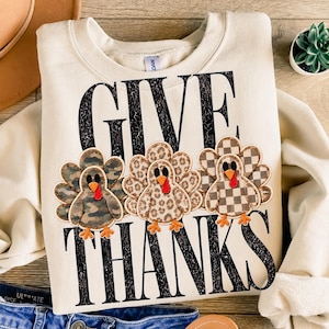 May include: A cream-colored crewneck sweatshirt featuring the black distressed text GIVE THANKS. Between the words are three turkey appliques: one with a camouflage pattern, one with a leopard print, and one with a brown and white checkerboard pattern.