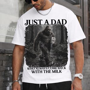 May include: A white short-sleeved t-shirt featuring a black and white graphic of a Bigfoot creature walking through a forest while holding a milk carton. The text above reads JUST A DAD and the text below reads WHO ALWAYS COME BACK WITH THE MILK.