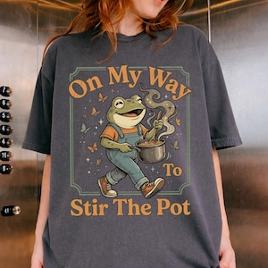 May include: Dark gray t-shirt featuring a cartoon frog wearing overalls and holding a pot with steam rising. The text reads "On My Way To Stir The Pot" in orange and white. Butterflies surround the frog, creating a whimsical design.