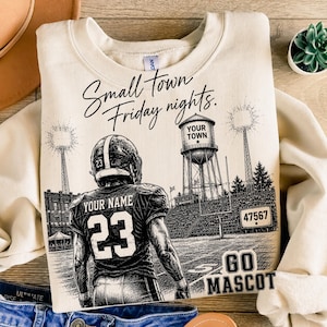 Op de afbeelding: Een crèmekleurige sweater met ronde hals en een zwarte afbeelding van een footballspeler in een shirt met nummer 23 en de tekst YOUR NAME. Het ontwerp bevat een watertoren met YOUR TOWN, stadionverlichting en de tekst Small town Friday nights en GO MASCOT.