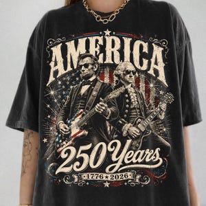 May include: Black t-shirt featuring an illustration of Abraham Lincoln and George Washington playing guitars. The word "AMERICA" is at the top, with "250 Years 1776-2026" below the figures. The design incorporates red, white, and blue elements.