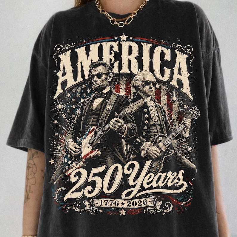 May include: Black t-shirt featuring an illustration of Abraham Lincoln and George Washington playing guitars. The word "AMERICA" is at the top, with "250 Years 1776-2026" below the figures. The design incorporates red, white, and blue elements.
