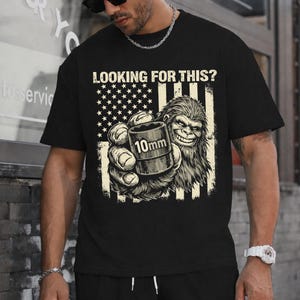 Könnte beinhalten: Schwarzes T-Shirt mit einer Grafik eines Bigfoot, der einen 10-mm-Steckschlüssel vor einem abgenutzten amerikanischen Flaggen-Design hält. Der Text "LOOKING FOR THIS?" befindet sich über der Grafik. Das Design ist in Grau- und Weißtönen gehalten.