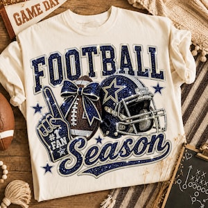 Pode incluir: Uma t-shirt cor de creme com um gráfico que apresenta o texto FOOTBALL SEASON em glitter azul escuro. O design inclui uma bola de futebol americano castanha com um laço azul, um capacete azul com uma estrela e um dedo de espuma azul #1 FAN.