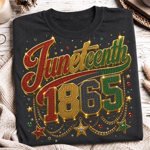 May include: Black t-shirt with a colorful, jeweled design that reads "Juneteenth 1865." The text is in red, green, and gold, with decorative stars and chains. The shirt is folded on a light-colored surface.
