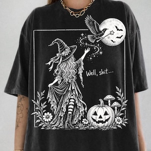 May include: A charcoal gray t-shirt featuring a white graphic print of a witch in a pointed hat reaching toward a flying crow and a full moon. The design includes a jack-o'-lantern, mushrooms, flowers, and the text "Well, shit..." in a script font.