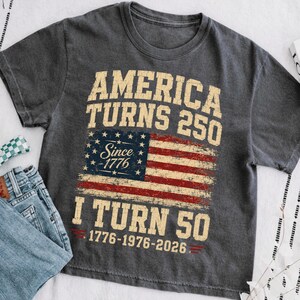 May include: Dark gray t-shirt with the text "America Turns 250" above a distressed American flag graphic. Below the flag, the text reads "I Turn 50" with the years 1776-1976-2026. The shirt is paired with blue jeans.