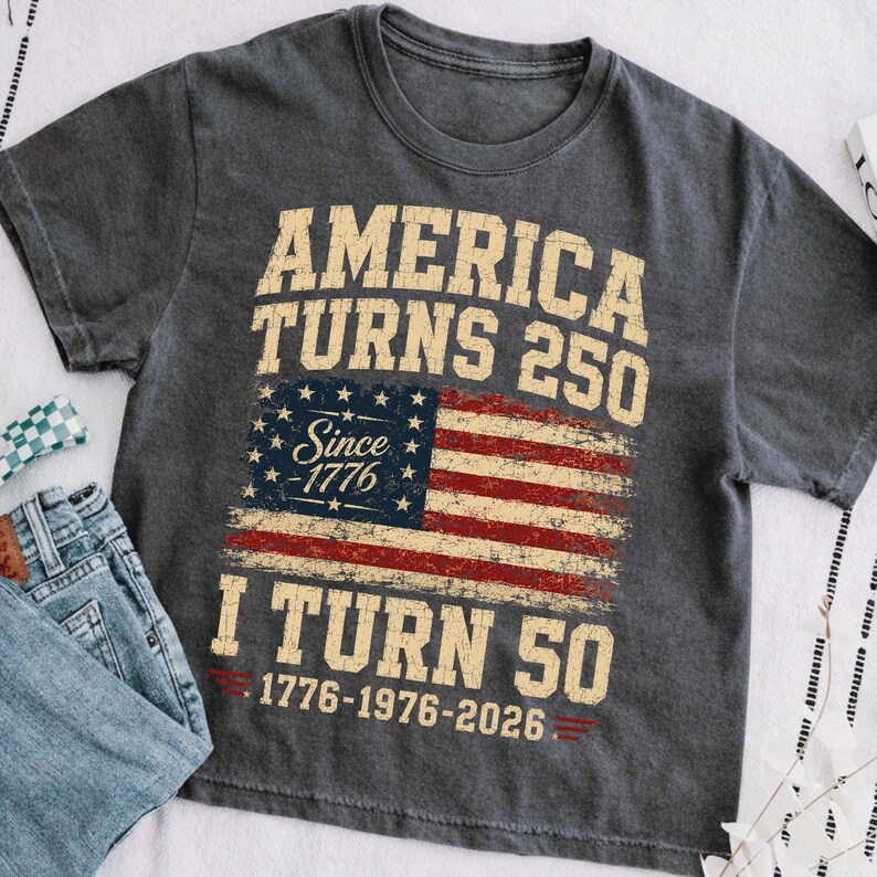May include: Dark gray t-shirt with the text "America Turns 250" above a distressed American flag graphic. Below the flag, the text reads "I Turn 50" with the years 1776-1976-2026. The shirt is paired with blue jeans.