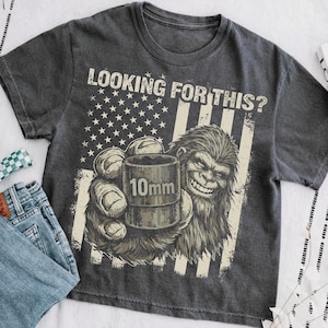 Könnte beinhalten: Dunkelgraues T-Shirt mit dem Text "LOOKING FOR THIS?" über einer Grafik von Bigfoot, der eine 10-mm-Steckschlüssel hält. Das Design enthält ein amerikanisches Flaggenmotiv mit Sternen und Streifen.