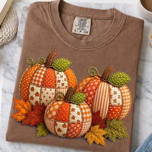 Op de afbeelding: Een bruin t-shirt met korte mouwen en een afbeelding van drie pompoenen gemaakt van diverse oranje, witte en groene stoffen patronen. De pompoenen zijn versierd met groene stoffen bladeren en oranje en gele herfstbladeren.