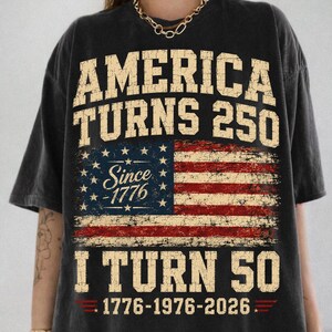 May include: Dark gray t-shirt with a distressed American flag design. The shirt features the text "AMERICA TURNS 250" above the flag and "I TURN 50" below, with the years 1776-1976-2026 at the bottom.