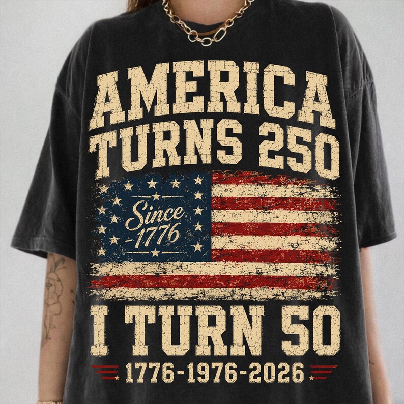 May include: Dark gray t-shirt with a distressed American flag design. The shirt features the text "AMERICA TURNS 250" above the flag and "I TURN 50" below, with the years 1776-1976-2026 at the bottom.